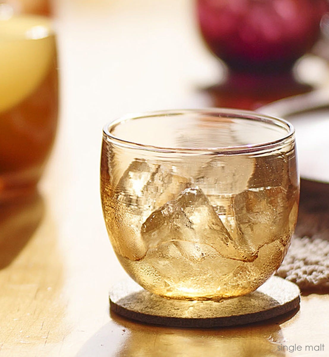 single malt drinker, transparent light amber hand-blown drinking glass filled with ice cubes and liquid on a gray coaster on a wood table.
