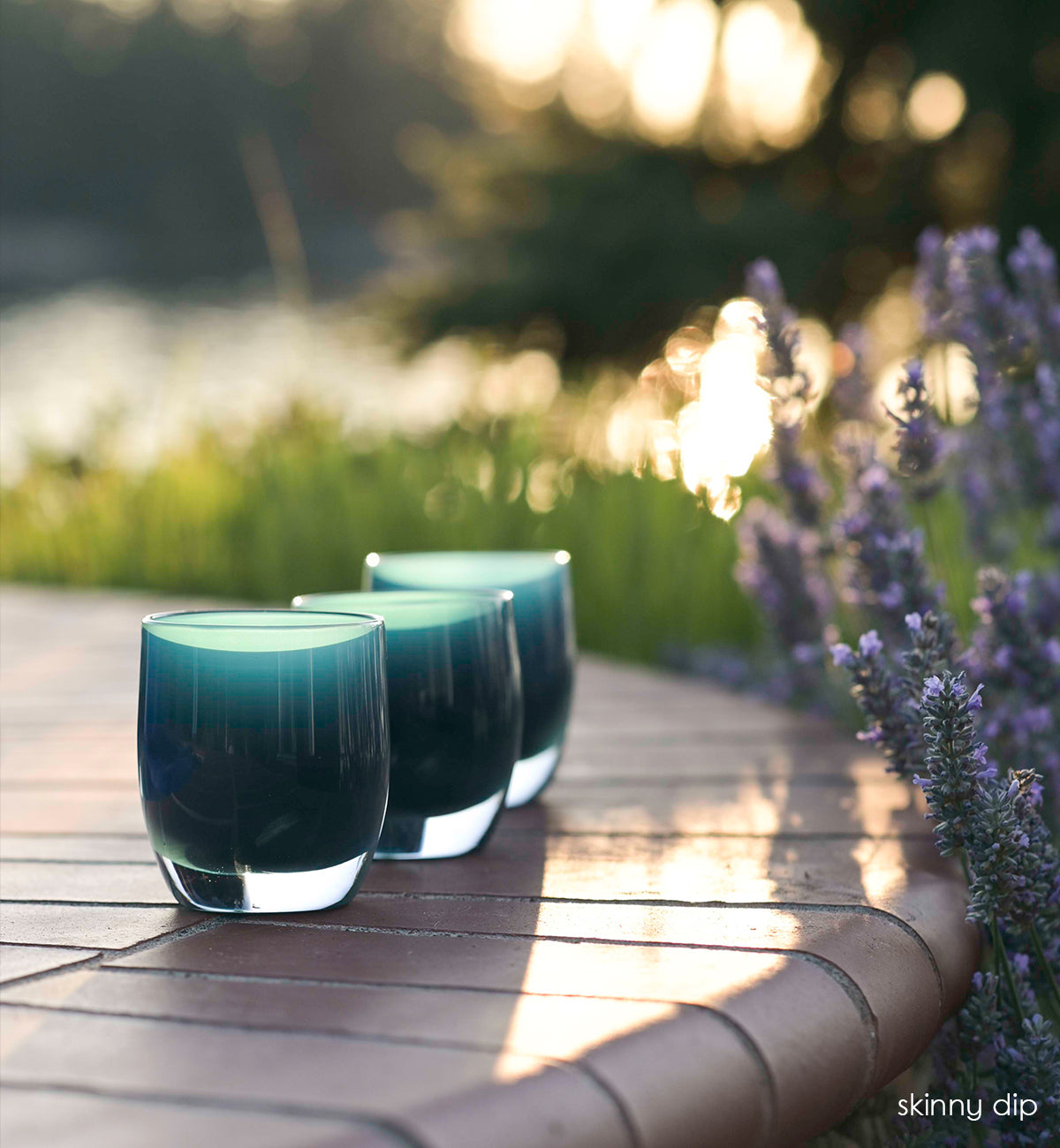 three skinny dip, dark teal hand-blown glass votive candle holders on a brick patio next to lavender.