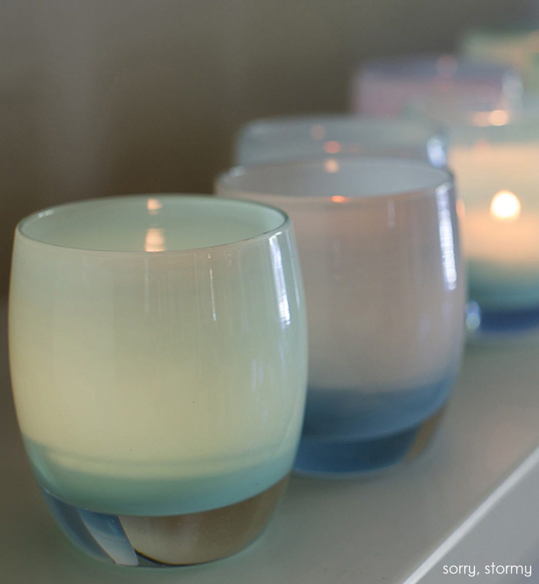sorry, light blue, hand-blown glass votive candle holder with stormy on cream surface.