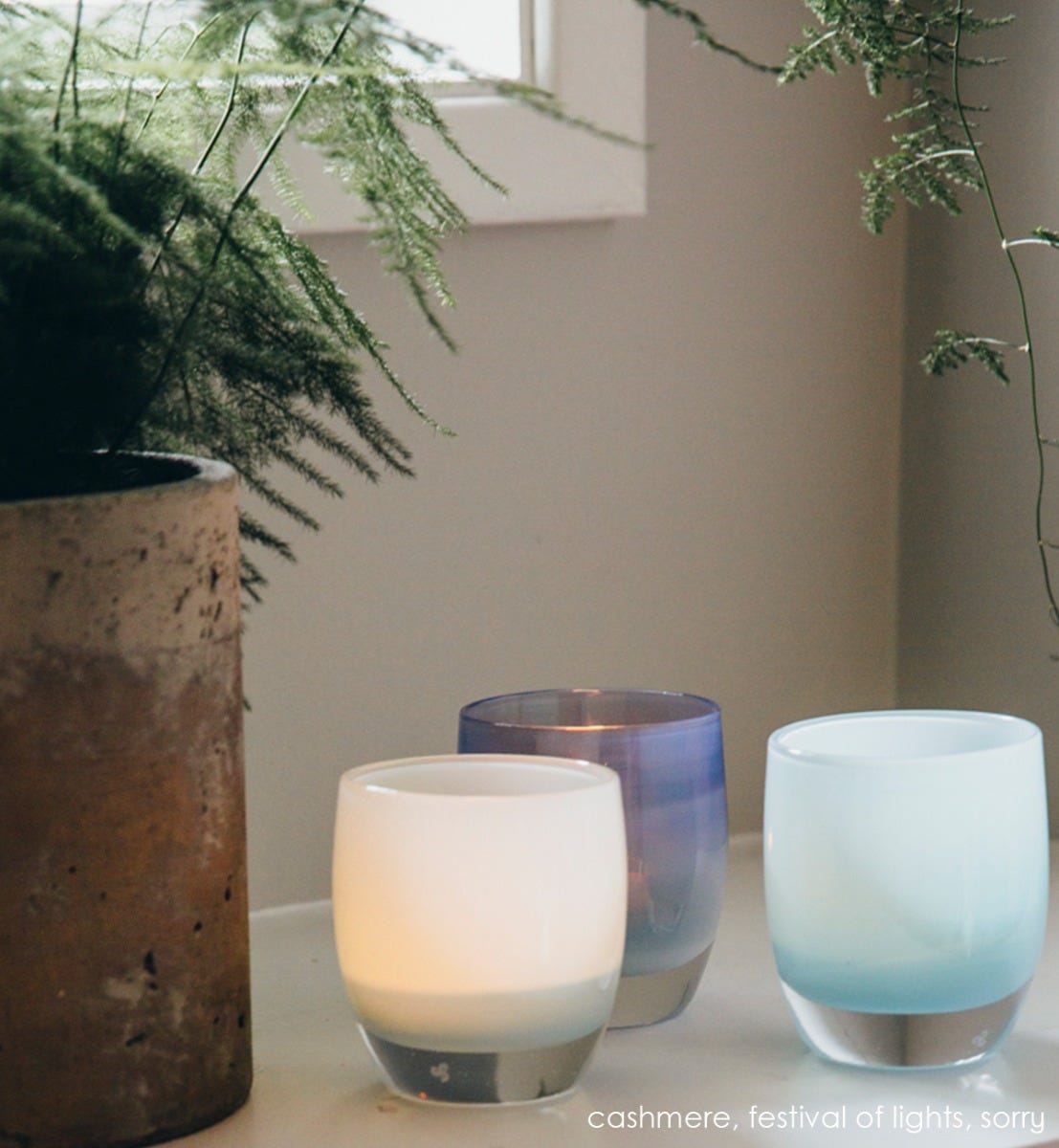 sorry, light blue, hand-blown glass votive candle holder. Paired with cashmere and festival of lights on cream table with greenery on both sides.