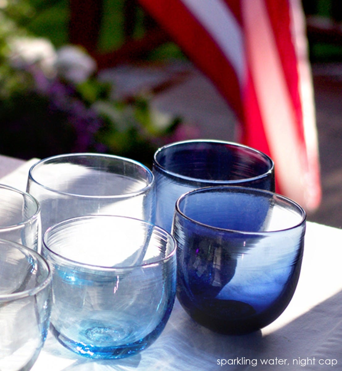 two sparking water drinkers, transparent light sky blue, hand-blown drinking glasses with night cap drinkers on an outdoor table with American flag in background.