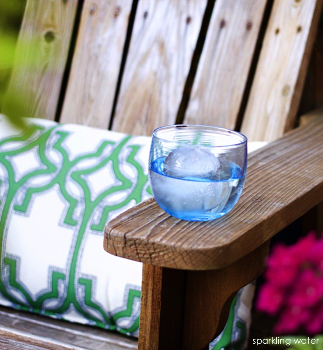 sparking water drinker, transparent light sky blue, hand-blown drinking glass with liquid and ice on a wood patio chair next to green patterned pillow.
