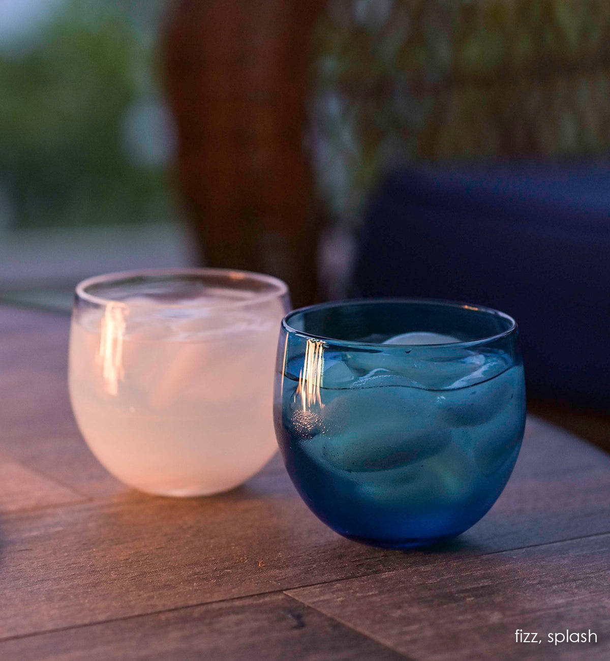 splash, hand-blown blue drinking glass next to fizz drinker, both filled with ice and liquid on an outdoor patio.