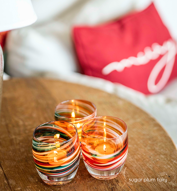 three sugar plumfairy, holiday multicolor textured, hand-made glass votive candle holders on a wood living room table next to a couch with festive pillows.