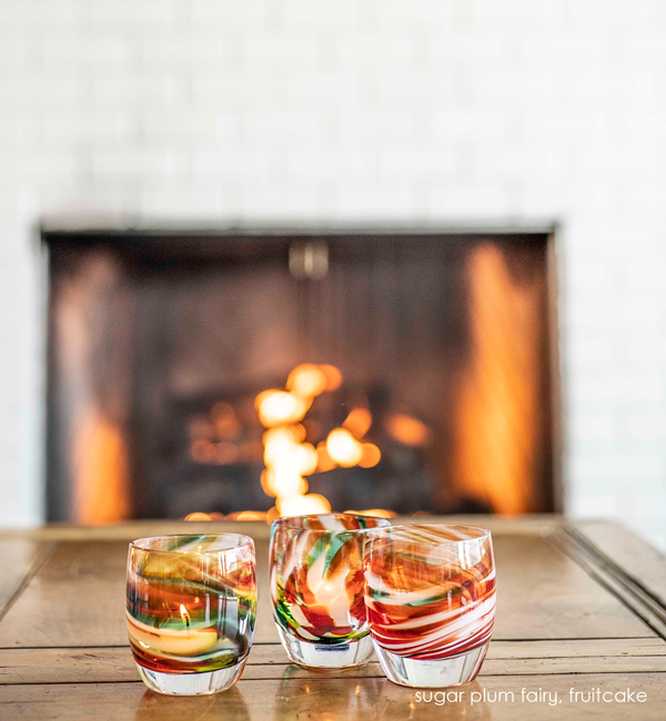 sugar plum fairy, holiday multicolor textured, hand-made glass votive candle holders with fruitcake on a wood table in front of a in use fireplace.