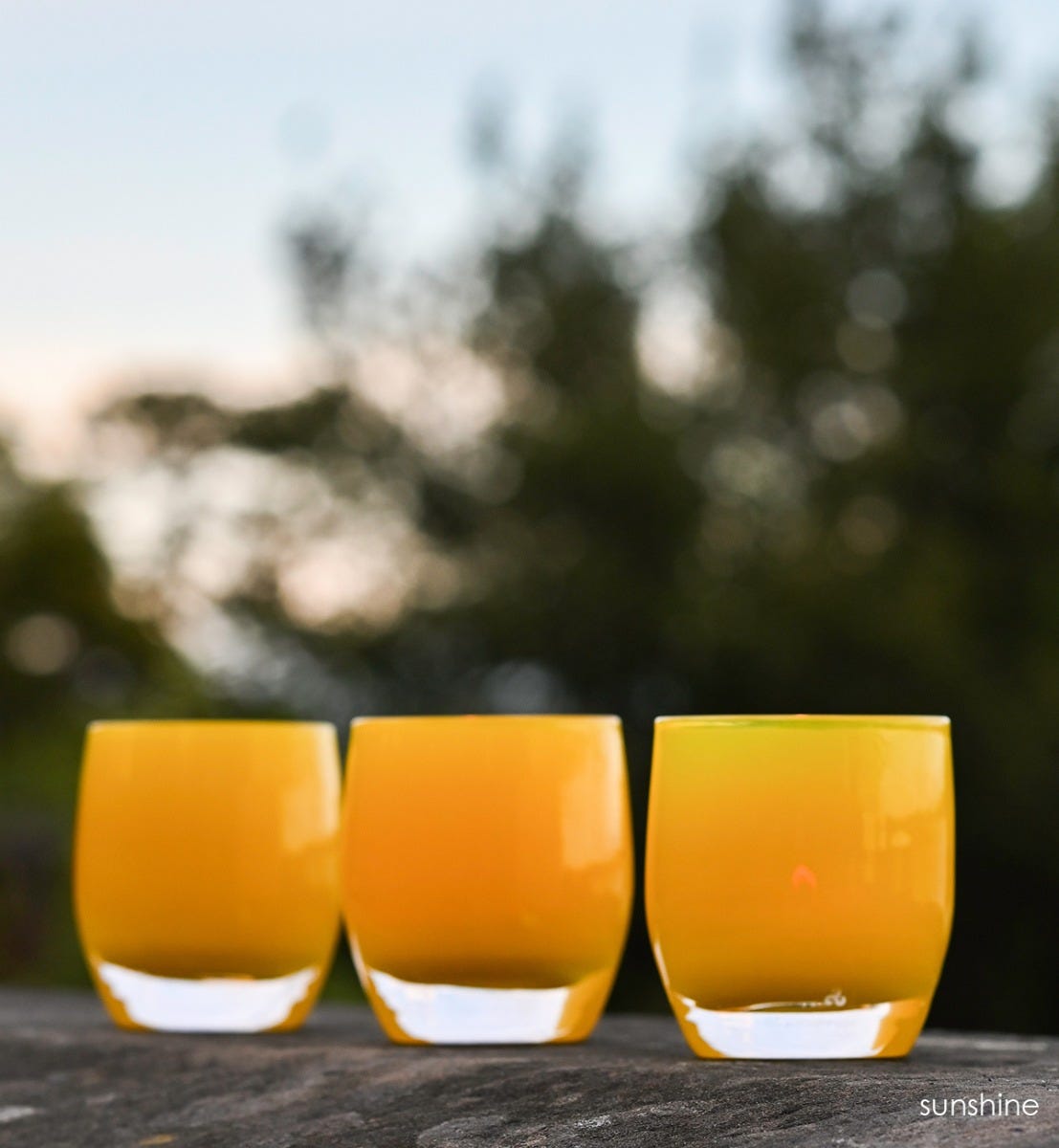three sunshine, opaque hand-blown bright yellow glass votive candle holders with blurred trees in background.
