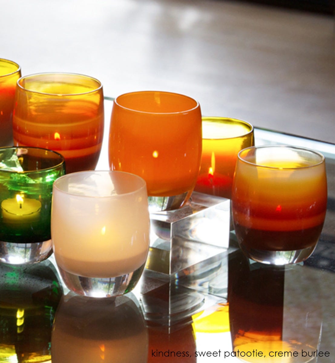 sweet patootie, apricot orange, hand-blown glass votive candle holder, with kindness and creme brûlée on a reflective surface and acrylic baby stand.