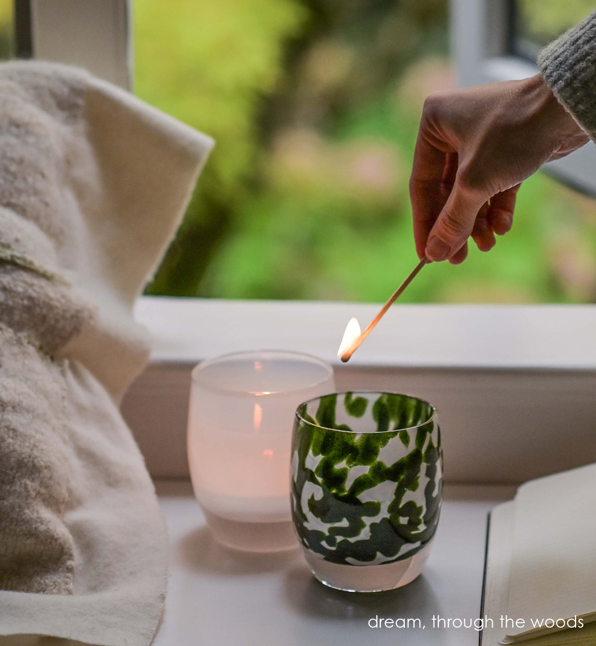 through the woods hand-blown dark green with glitter glass votive candle holder with a lit match above it, window behind and next to a pillow, book, and dream..