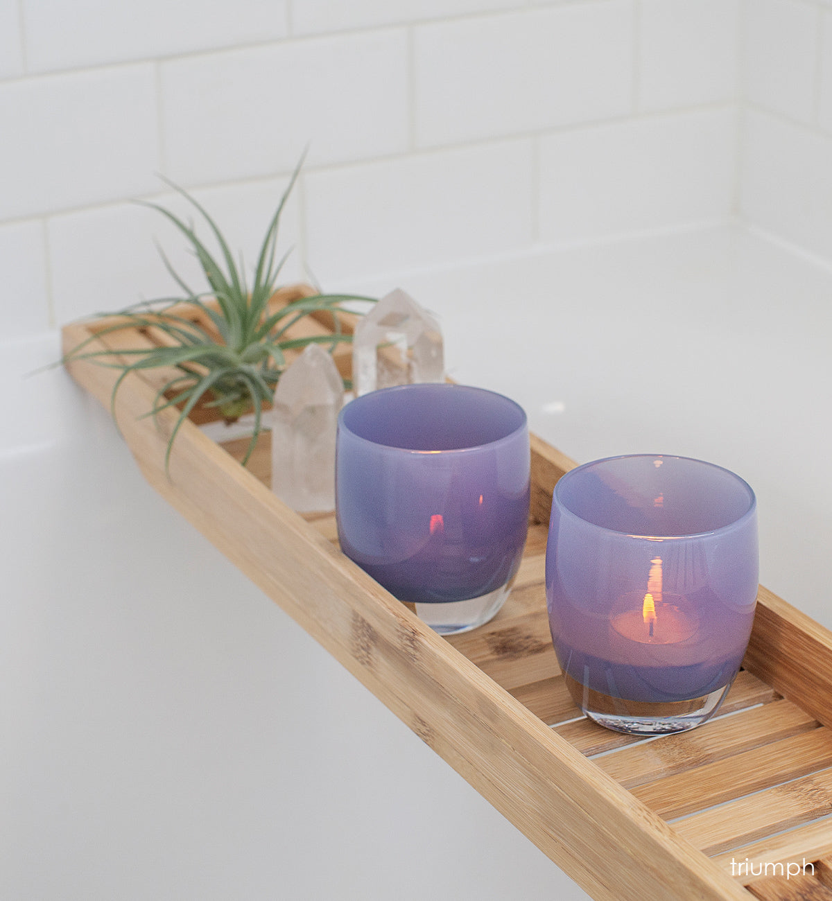 two triumph, misty purple, hand-blown glass votive candle holders on a wood bath tray next to crystals and a plant.