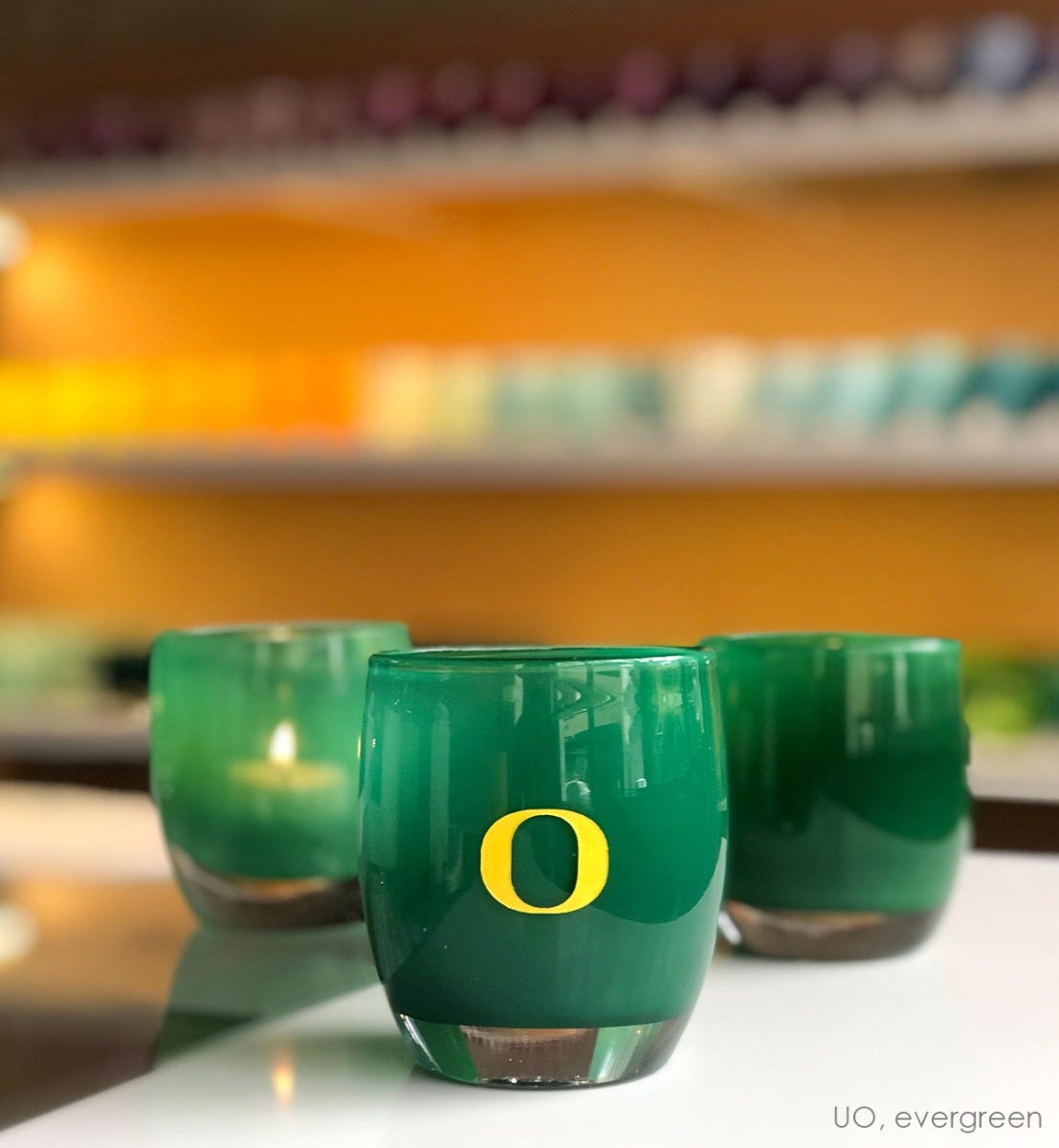 UO, green with sandblasted University of Oregon etching hand painted in yellow, hand-blown glass votive candle holder. Paired with evergreen on white and clear store table.