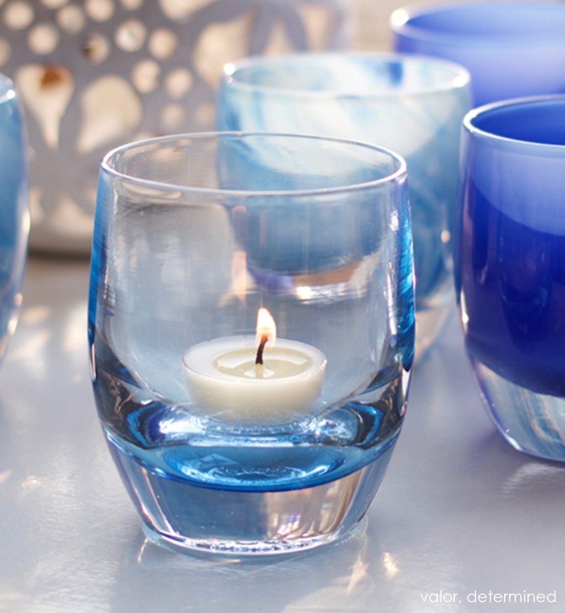 valor clear light blue hand-blown glass votive candle holder with a tealight lit on a white table with determined.