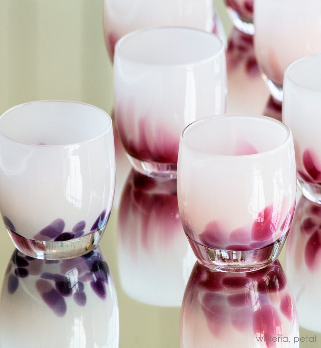 wisteria, white with purple petals emerging from the bottom, hand-blown glass votive candle holder, paired with petal on a reflective surface.