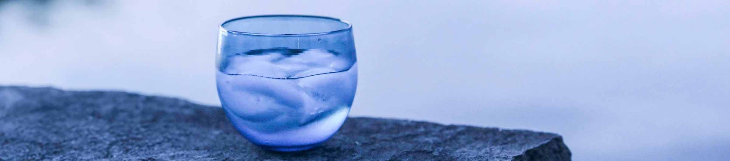 transparent blue splash drinker filled with ice water, sits on rock over looking calm water.