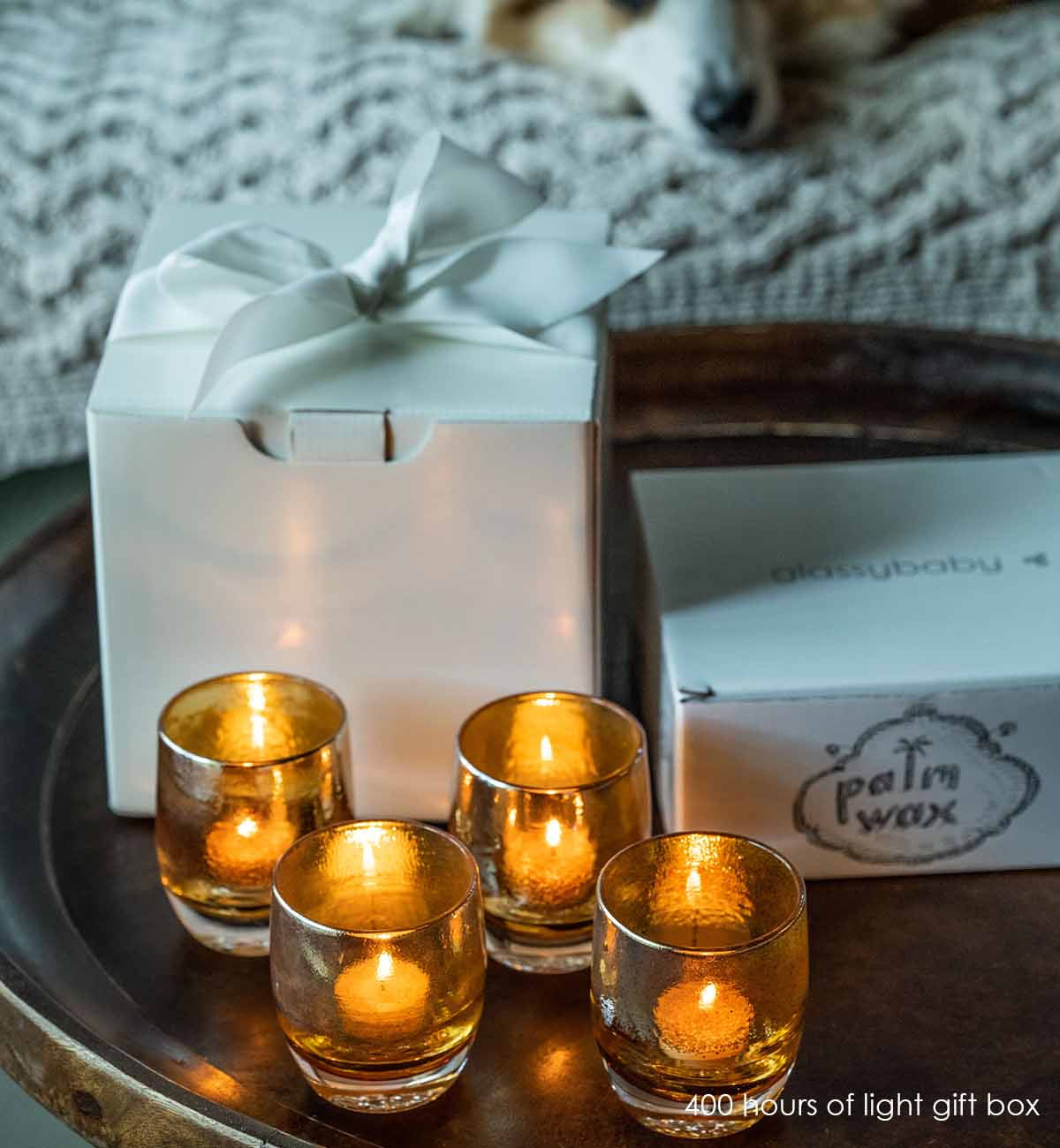 four hundred hours gift box, one hundred palm wax tea lights, and four stay gold hand-blown glass votives candle holders with bowed white box on a wood side table with couch and dog in background.