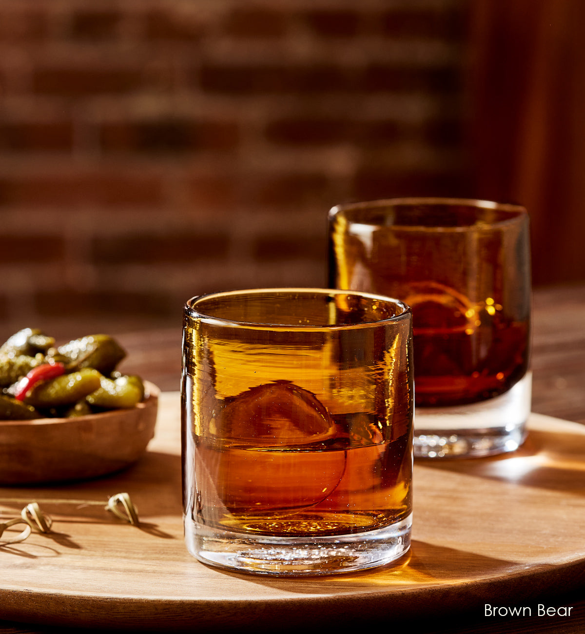 two Brown Bear rockers, dark brown transparent hand-blown glass lowball drinking glasses with liquid and ice, atop a wood cutting board next to pickles.