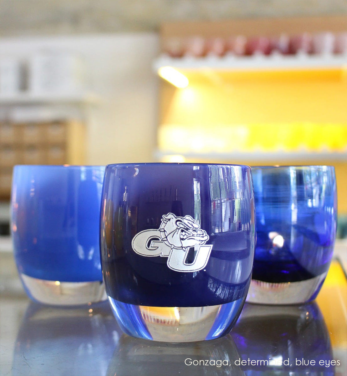 Gonzaga, Gonzaga University mascot logo in silver on navy blue, hand-blown glass votive candle holder. Paired with determined and blue eyes on a store counter.