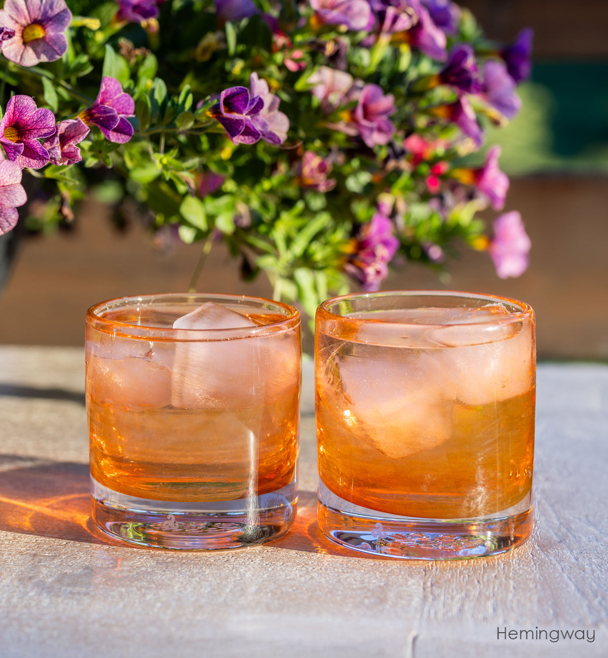 Hemingway coral rocker set - two coral hand-blown low ball whiskey glasses filled with water and ice. pink and purple flowers in the background.