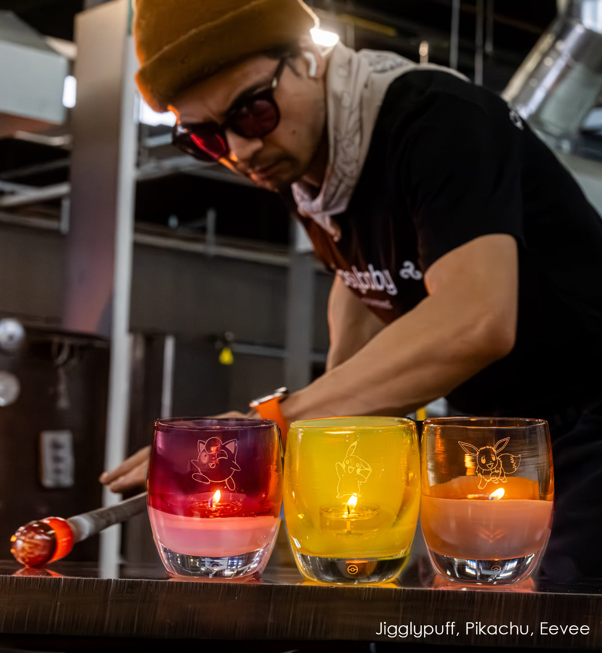 Person hand-blowing glass candle holders on a table, with 'Pokémon', Pikachu, Eevee, and Jigglypuff designs in hot shop.