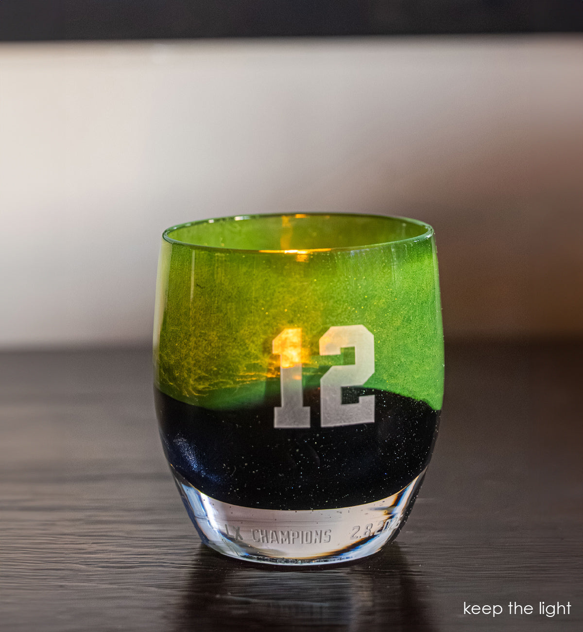 Close-up of a green and blue hand blown glass candle holder with '12' engraved on it on a wooden surface