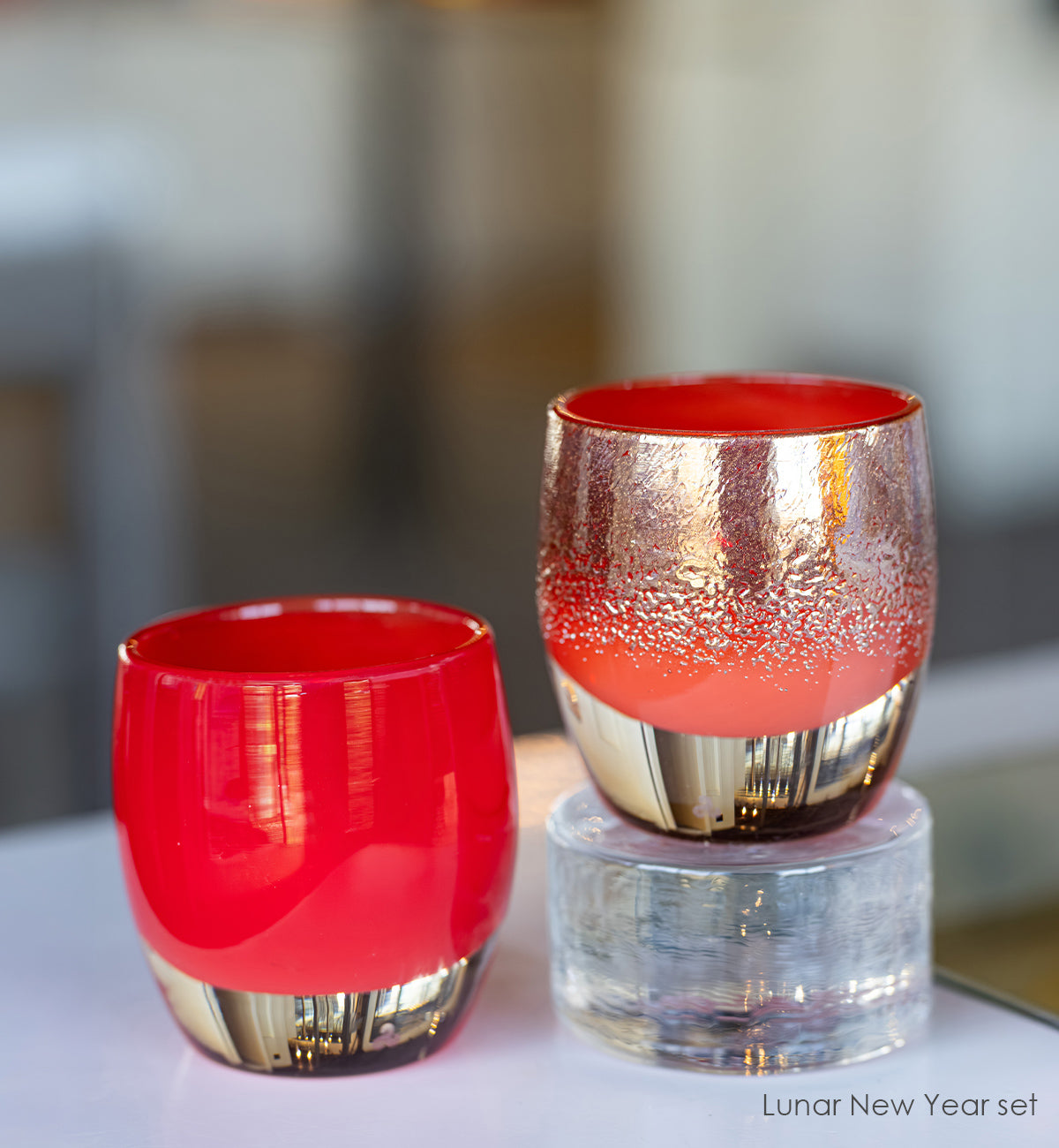 Two red and gold glass tumblers on a white surface with a blurred background.
