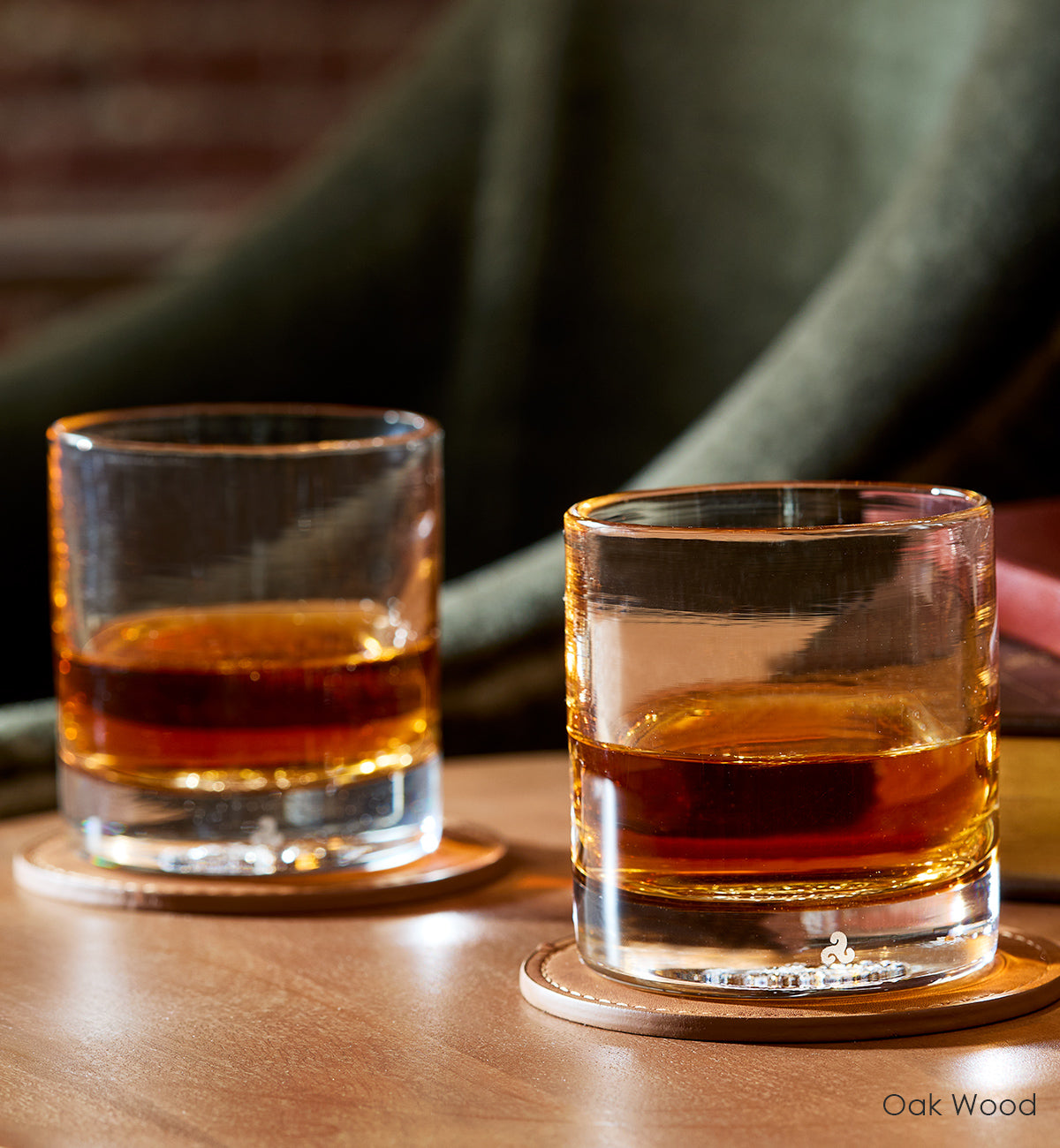 two Oak Wood rockers, light brown hand-blown glass lowball drinking glasses. with liquid inside on leather coasters atop a wood table with green chair in background.