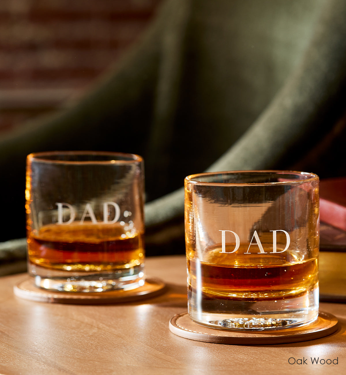 two Oak Wood rockers, light brown hand-blown glass lowball drinking glasses. with the word dad custom etched on two leather coasters atop a wood table with green chair in background.