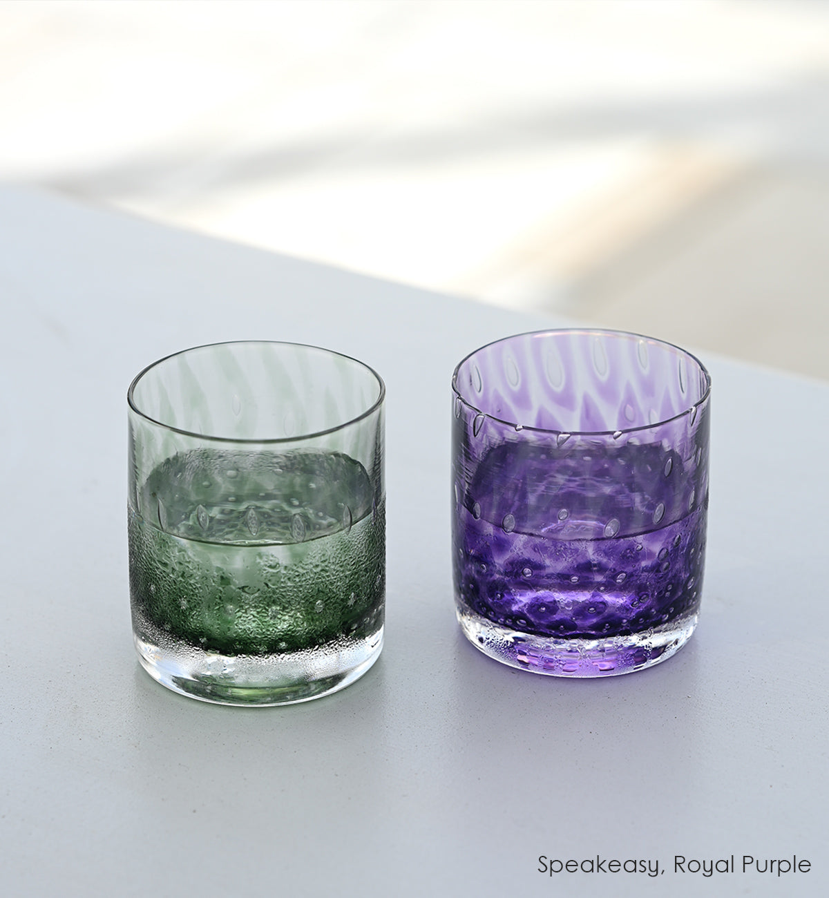 Speakeasy and Royal Purple rocker, dark green and purple hand-blown lowball glasses with bubble patterns sitting atop white surface.