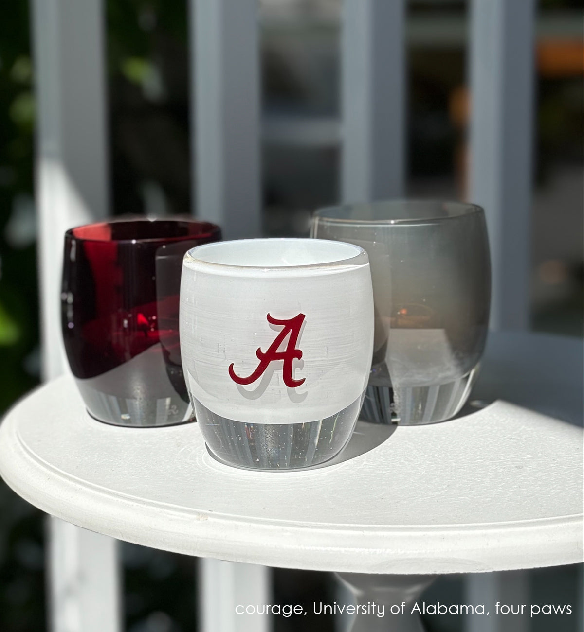 Alabama, etched University of Alabama college logo on a white hand-blown glass candle holder sitting on a white table with a courage and four paws glassybaby.