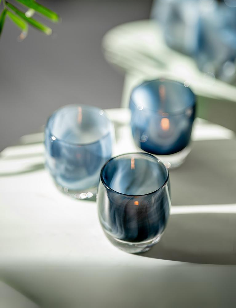 3 dark blue and white swirled patterned hand-blown glass candle holders names 'i see you' sitting on a white table in the sun.