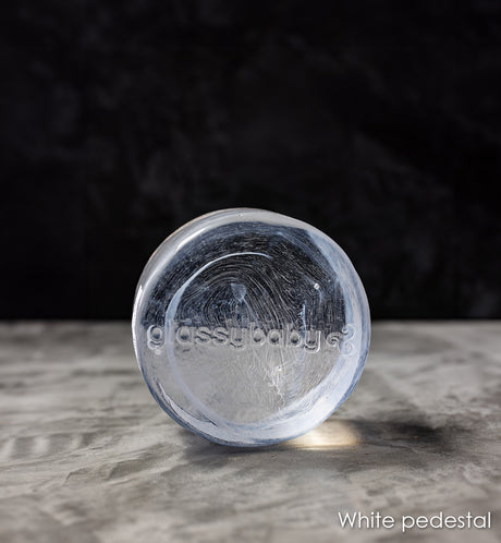 white transparent glass pedestal with 'glassybaby' branding on a gray surface with a dark background
