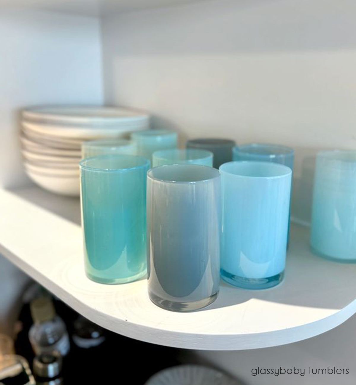 several glassybaby tumblers. aqua, grey, turquoise and blue hand-blown water glasses on white kitchen shelf next to cream ceramic bowls.