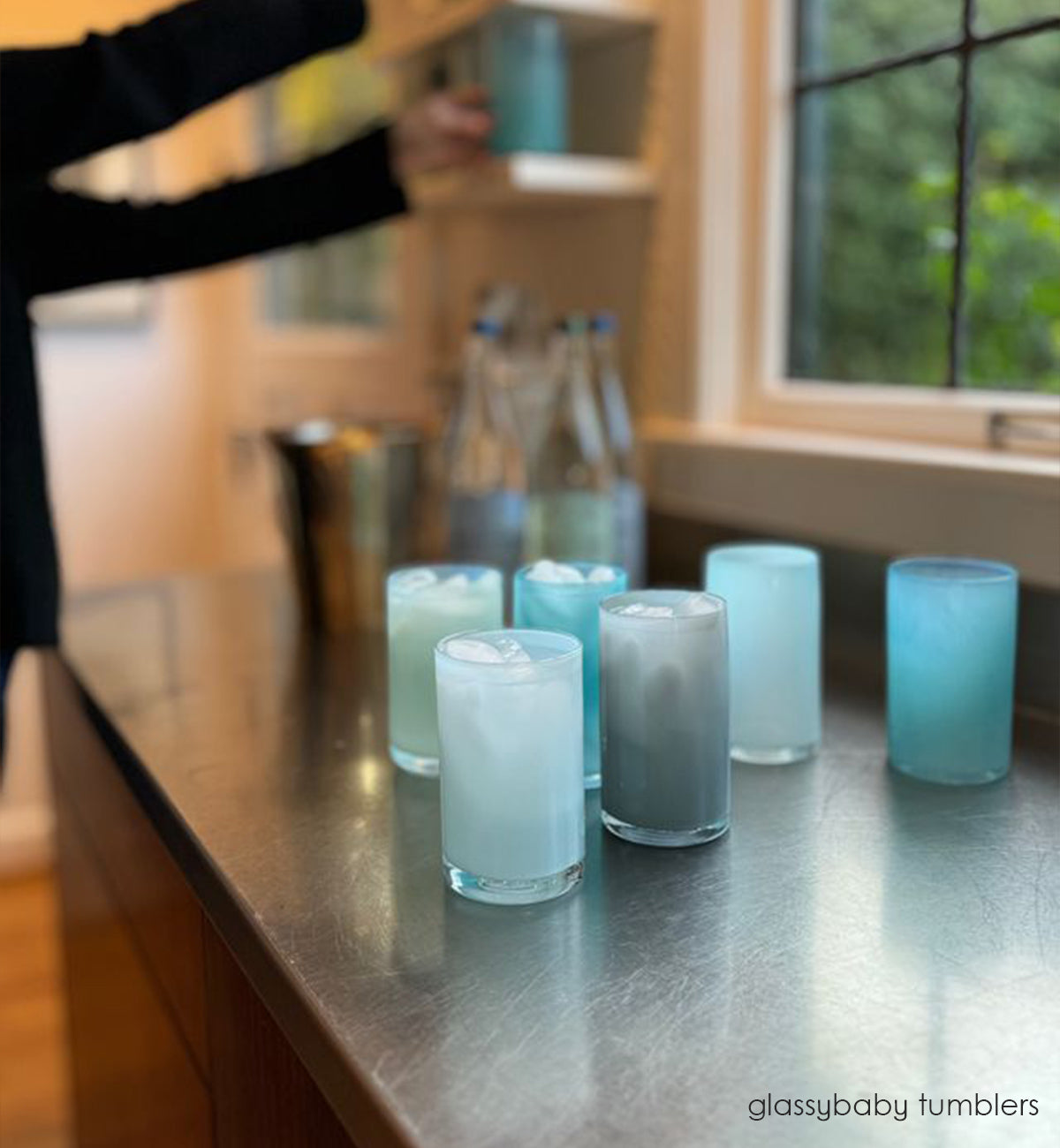 grey, blue, turquoise, and aqua tumblers, hand-blown water glasses on a kitchen counter with liquid and ice and with someone grabbing a turquoise tumbler off kitchen shelf in background.