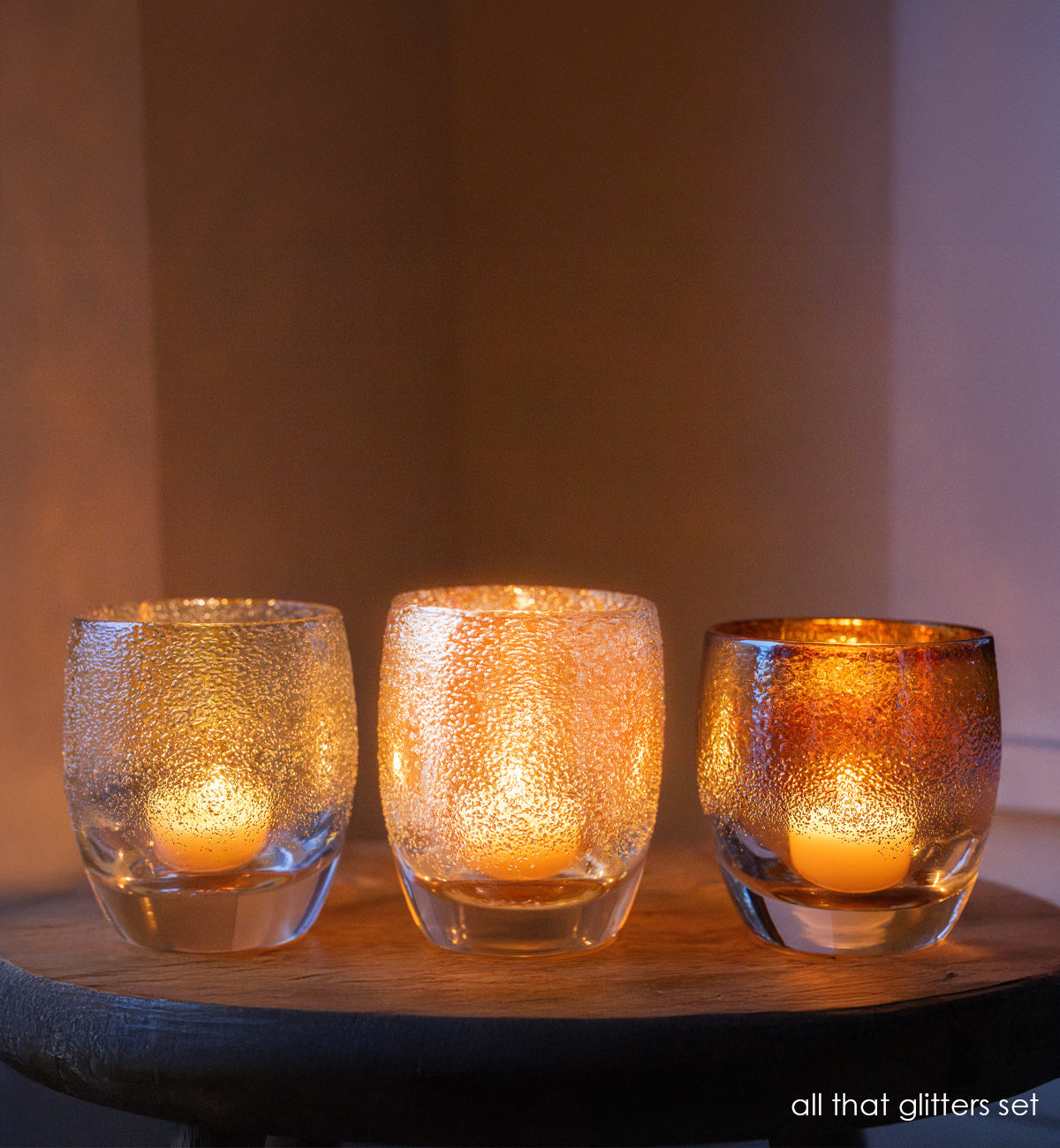sliver gold, white gold and yellow gold, hand-blown glass candle holders lit on a wood table. 