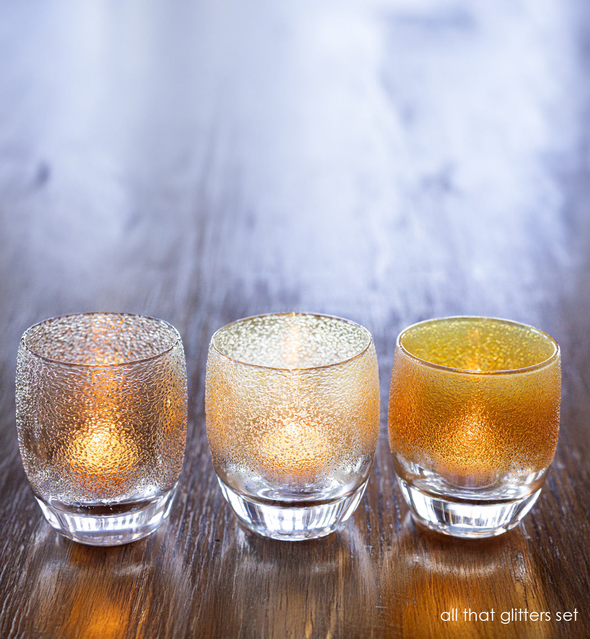 sliver gold, white gold and yellow gold, hand-blown glass candle holders lit on a wood table. 