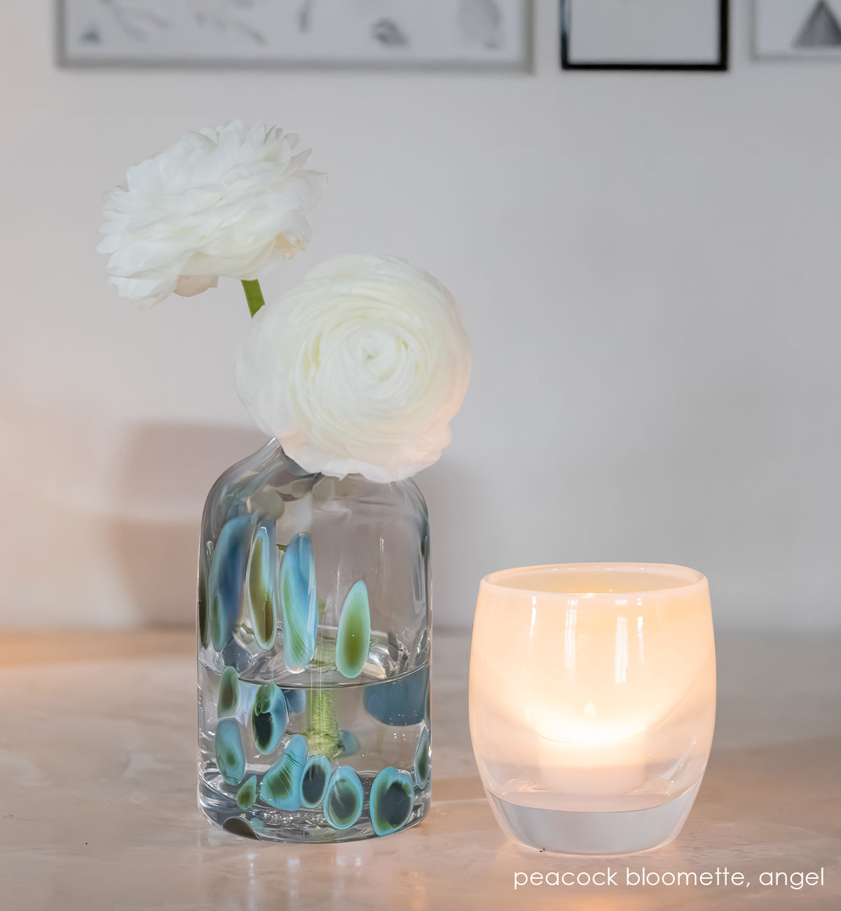 Glass bloomette vase with peacock design and white flowers, next to a lit white angel candle on a white surface.