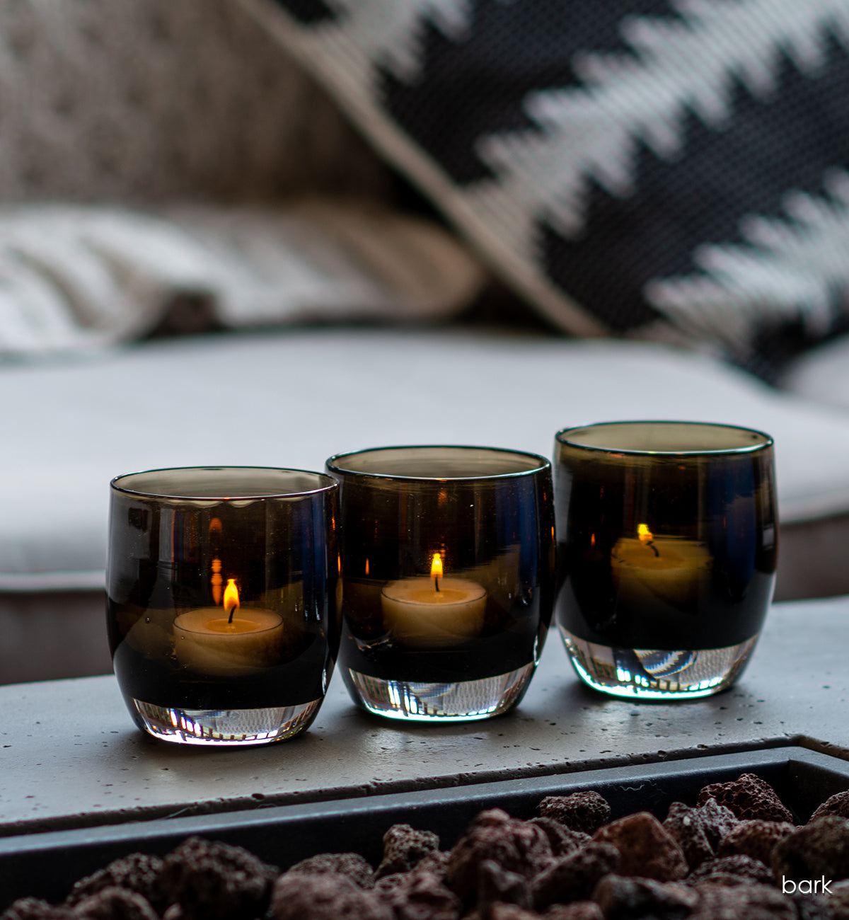 three 'bark', transparent rich dark brown, hand-blown glass votive candle holder by a outdoor fire place.