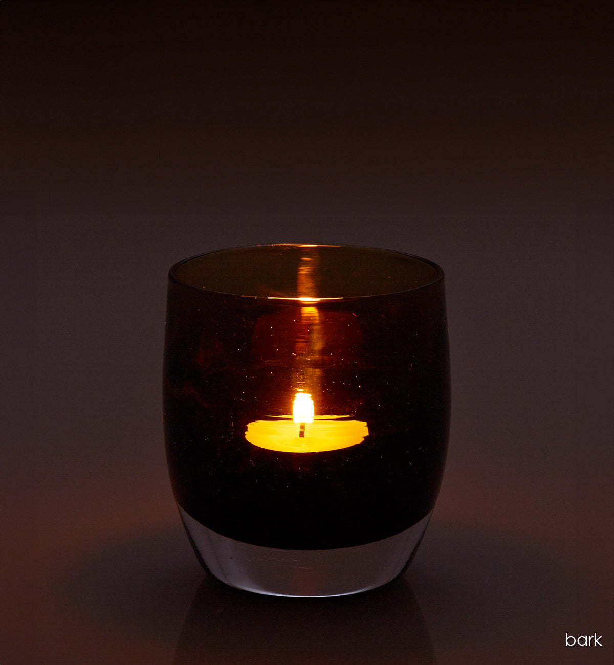 bark, transparent rich dark brown, hand-blown glass votive candle holder with a lit candle on a dark background