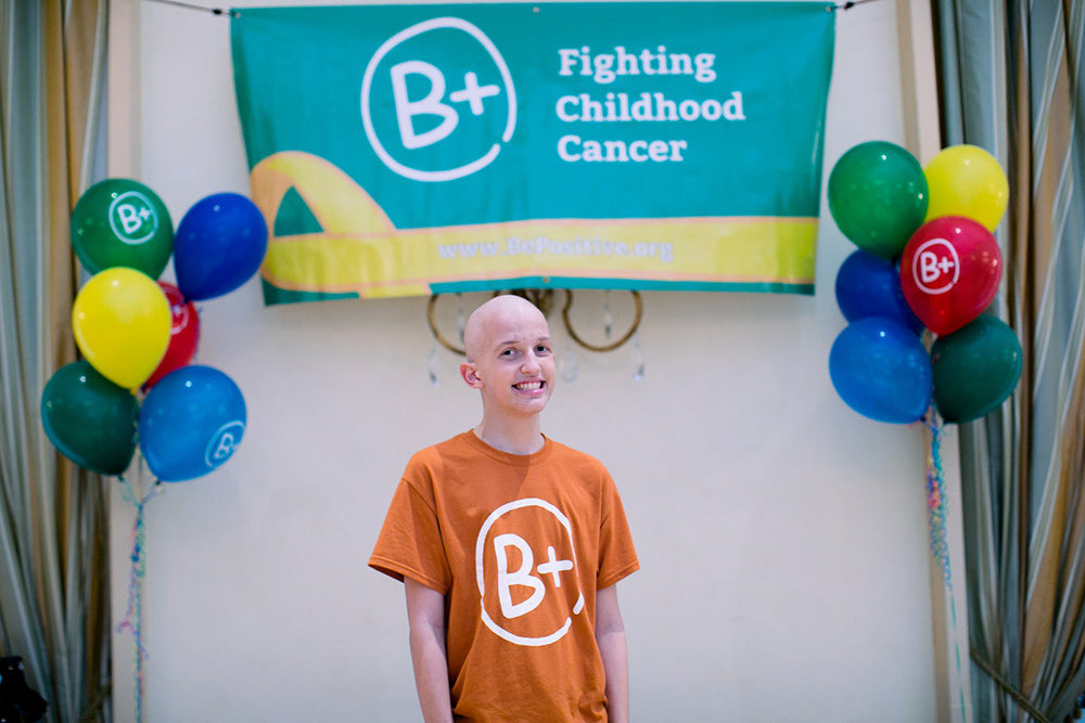 a young cancer patient in a B+ shirt with baloons and banner in background titled 'Fighting Childhood Cancer'