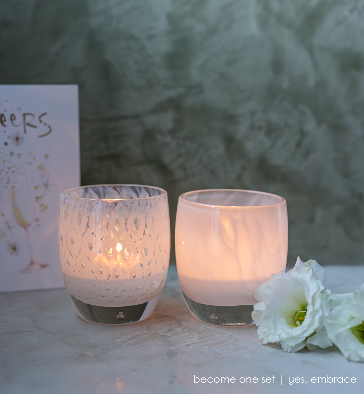 become one, set of 2 white handblown glass votive candle holders, yes and embrace lit with white flowers next to them.