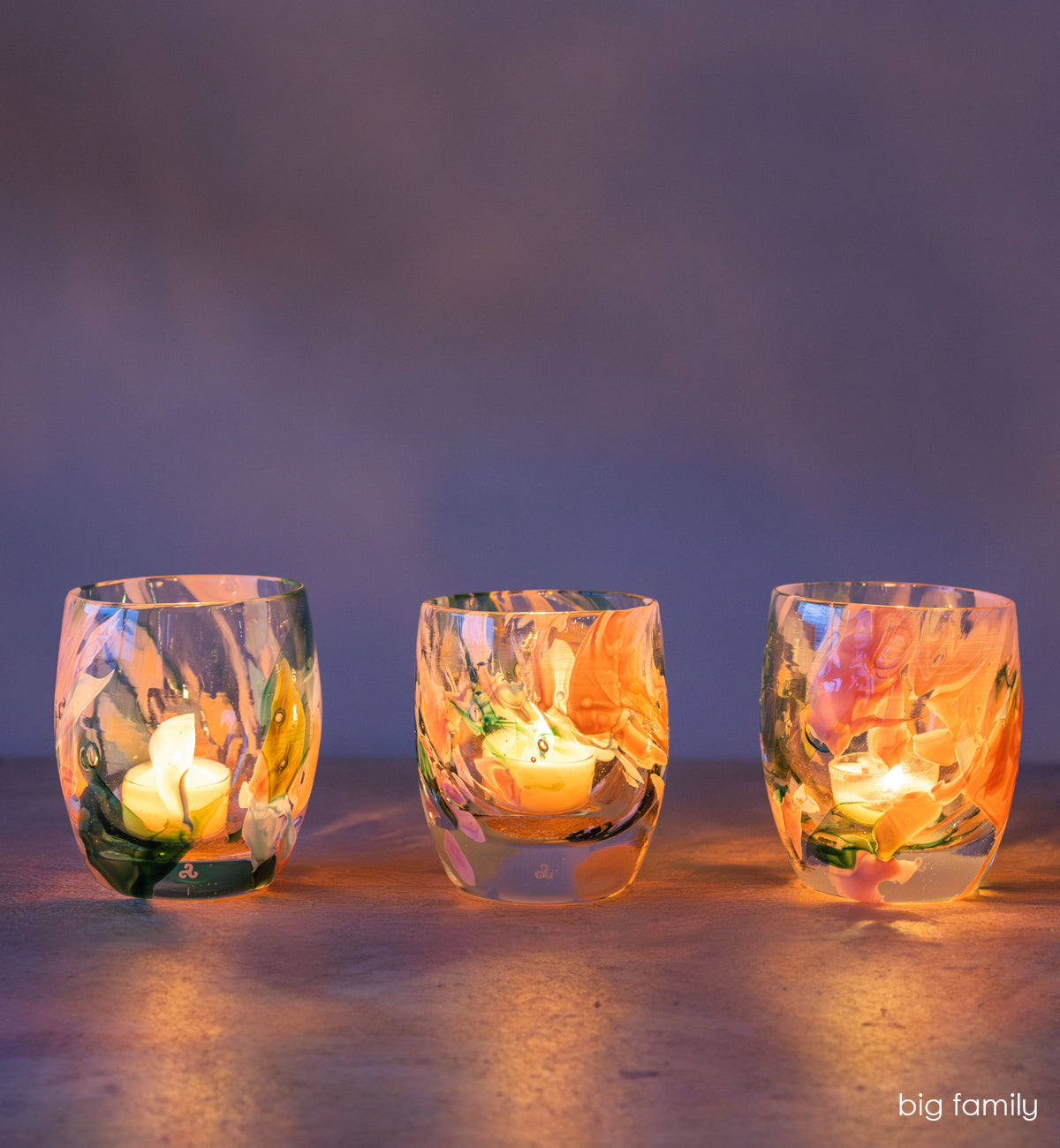 group of 'big family' multi-color (pinks, greens, light blues) hand-blown glass candle holder lit on gray surface. 