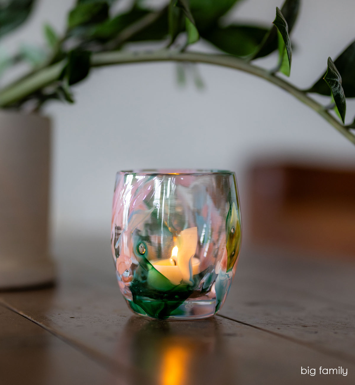 'big family' multi-color (pinks, greens, light blues) hand-blown glass candle holder lit on wood table with green plant hanging over it. 