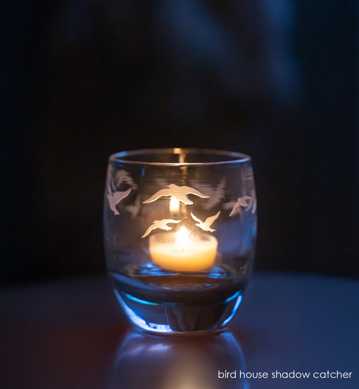 hand-blown glass candle holder with bird designs containing a lit candle on a dark background