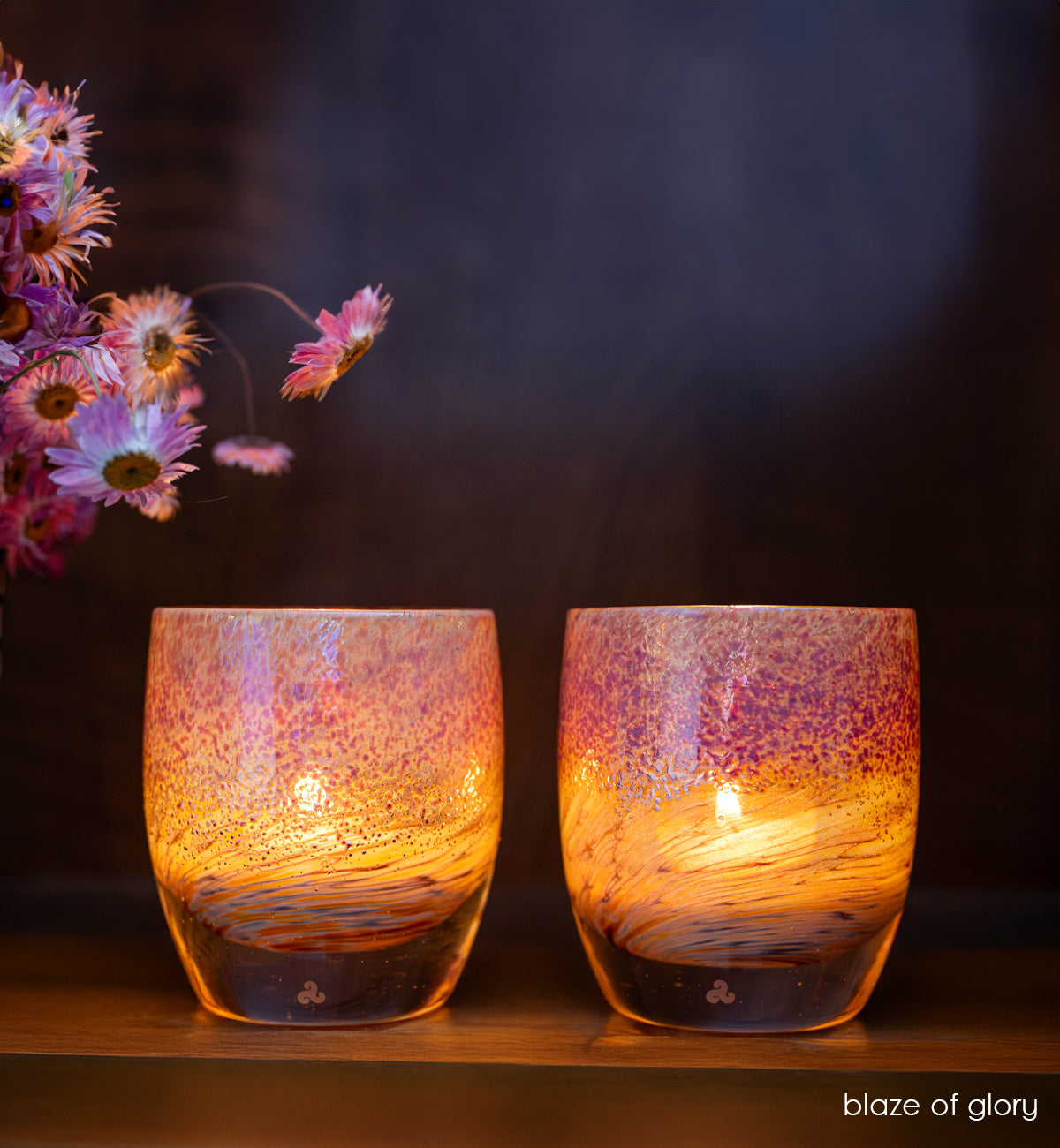 two blaze of glory, metallic hand-crafted glass votive candle holders on self with a black and white vase with pink flowers. 