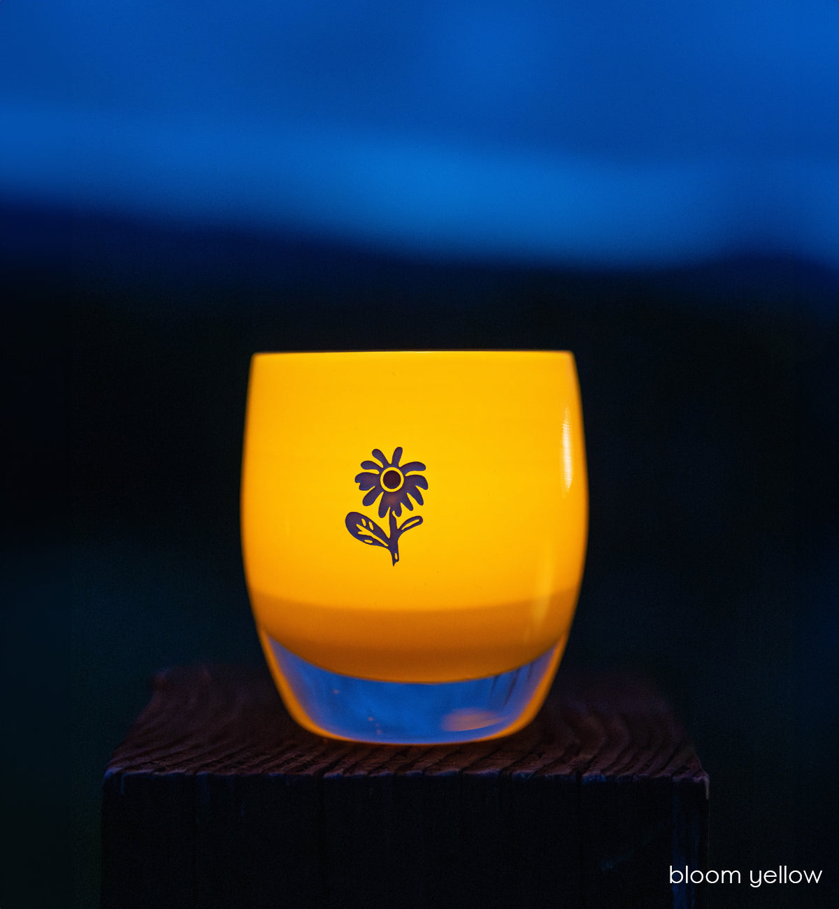 bloom yellow glassybaby with white flower etched on it sitting on wood shelf at night