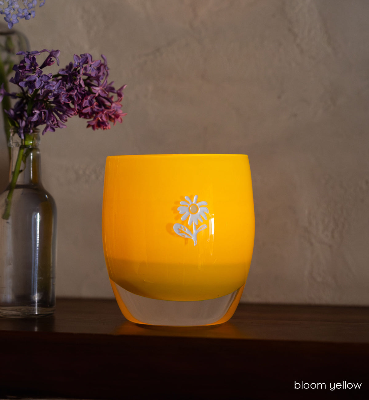 bloom yellow glassybaby with white flower etched on it sitting on wood shelf. 