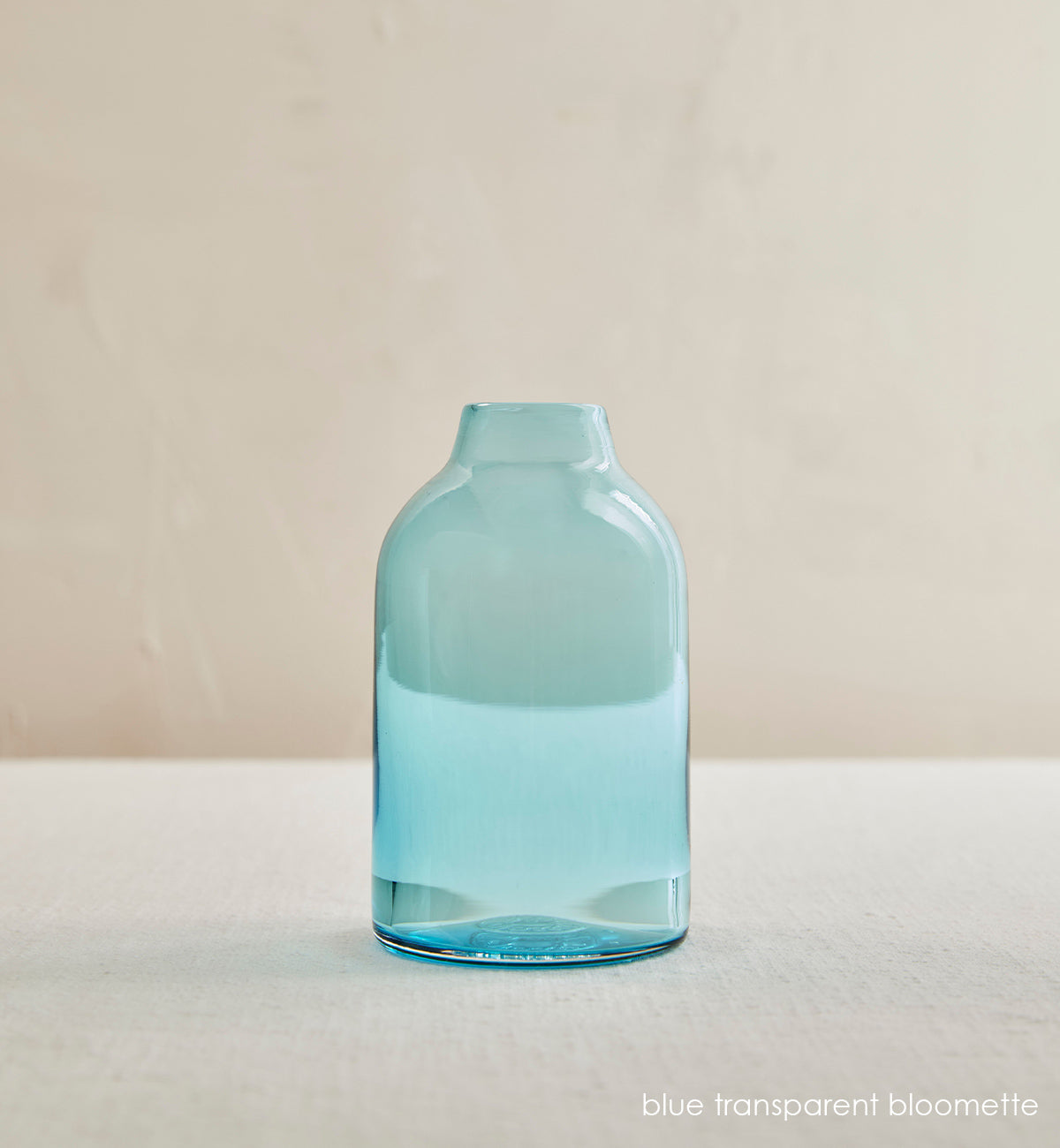 blue transparent bloomette, light transparent blue hand-blown glass flower bud vase in front of cream background.