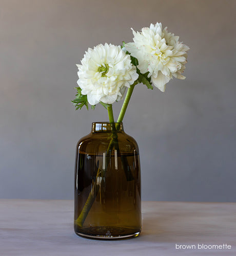  transparent brown hand-blown glass flower bud vase with white flowers on wood table and gray background