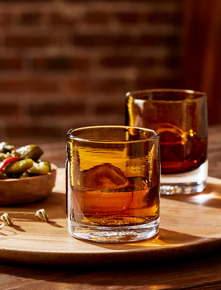 two Brown Bear, transparent rich dark brown, hand-blown lowball drinking glasses on a wood cutting board next to pickles.