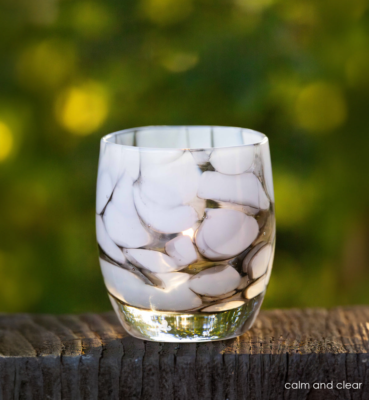calm and clear gray and white petal, hand-blown glass votive candle holder on wood rail with green trees in background.
