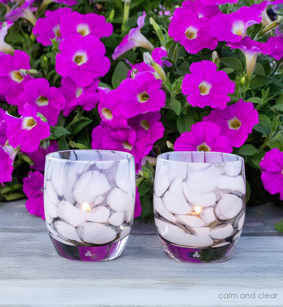 two calm and clear gray and white petal, hand-blown glass votive candle holders on gray table with purple flowers. 
